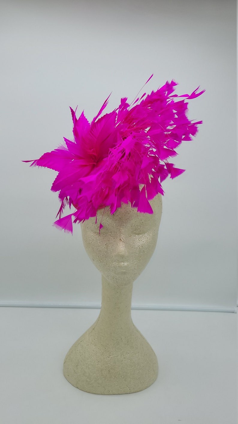 May include: A bright pink feather headpiece on a white mannequin head. The feathers are arranged in a circular shape and are very full.