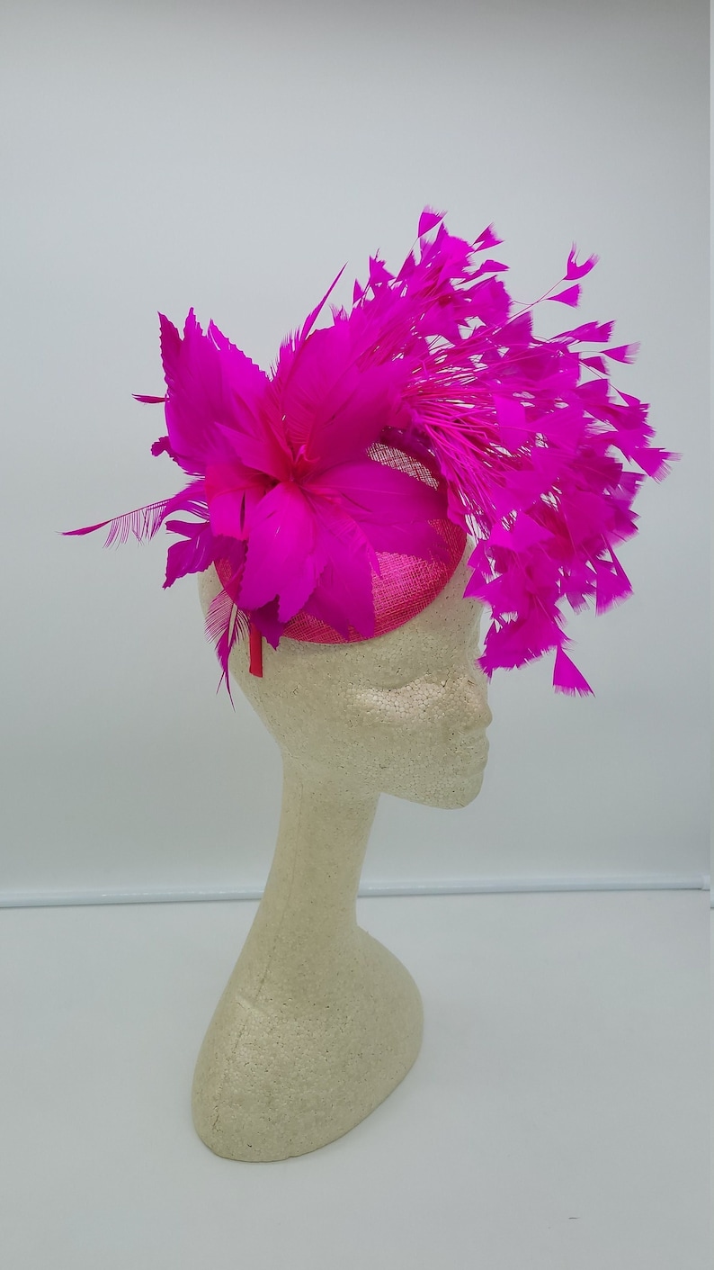 May include: A bright pink fascinator with a large feather design. The fascinator is made of a soft, lightweight material and is perfect for adding a touch of elegance to any outfit.