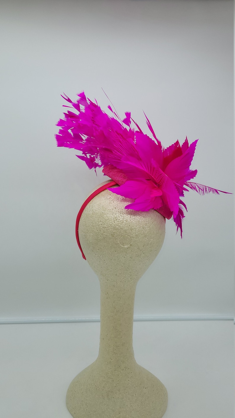 May include: A bright pink feather headband with a small, round base. The headband is displayed on a white mannequin head.