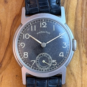 May include: A vintage Hamilton wristwatch with a black dial and silver hands. The watch features a small seconds subdial and is set in a silver case. It has a black leather strap.