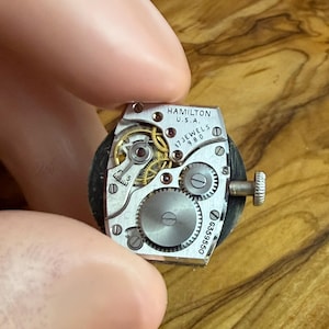 May include: Close-up of a vintage Hamilton watch movement. The silver mechanism features intricate gears, jewels, and the text "HAMILTON U.S.A. 17 JEWELS 980". A small crown is visible on the side. The watch movement is held between fingers.