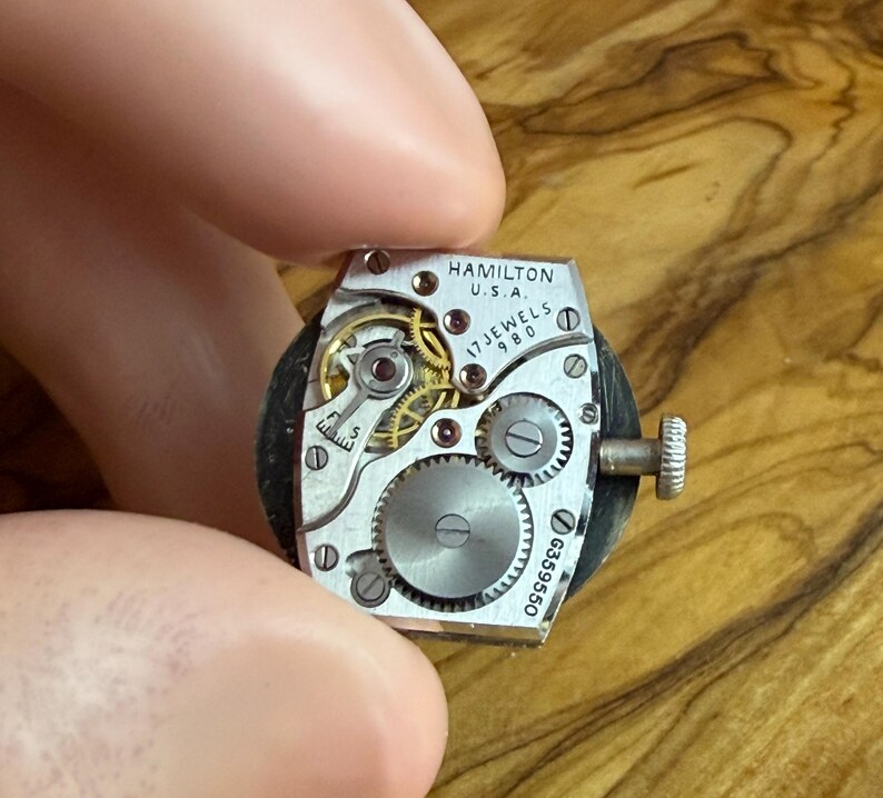 May include: Close-up of a vintage Hamilton watch movement. The silver mechanism features intricate gears, jewels, and the text "HAMILTON U.S.A. 17 JEWELS 980". A small crown is visible on the side. The watch movement is held between fingers.