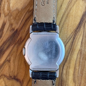 May include: A silver watch with a black leather strap. The watch has a round face and a stainless steel back. The strap is black, textured leather. The watch is on a wooden surface.
