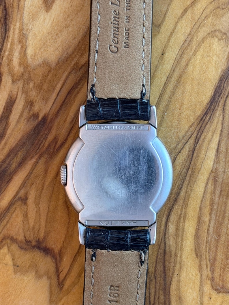 May include: A silver watch with a black leather strap. The watch has a round face and a stainless steel back. The strap is black, textured leather. The watch is on a wooden surface.