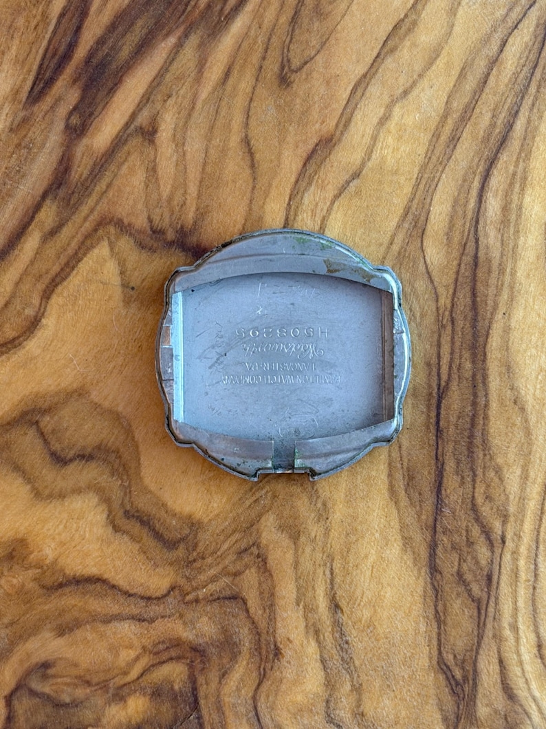 May include: A vintage silver-toned watch case with a rectangular interior. The case features a curved, decorative design around the edges. The back of the watch case has engraved text, including the number H508305.