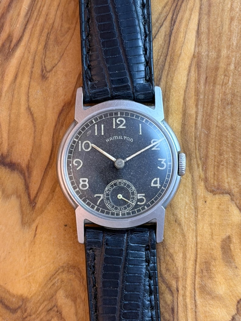 May include: A vintage Hamilton wristwatch with a black dial and silver hands. The watch features a small seconds subdial and is set in a silver case. It has a black leather strap.