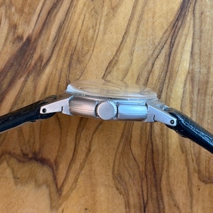 May include: A silver wristwatch with a black leather strap. The watch features a round face with a clear crystal and a silver crown. The side profile of the watch is visible against a wooden backdrop.