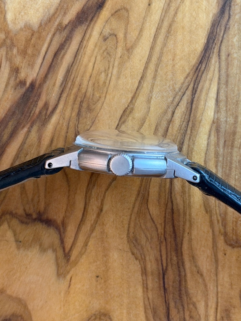 May include: A silver wristwatch with a black leather strap. The watch features a round face with a clear crystal and a silver crown. The side profile of the watch is visible against a wooden backdrop.