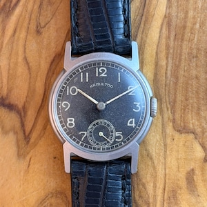 May include: A vintage Hamilton wristwatch with a black face, silver hands, and Arabic numerals. The watch has a small seconds dial and is set in a silver case with a black leather strap.