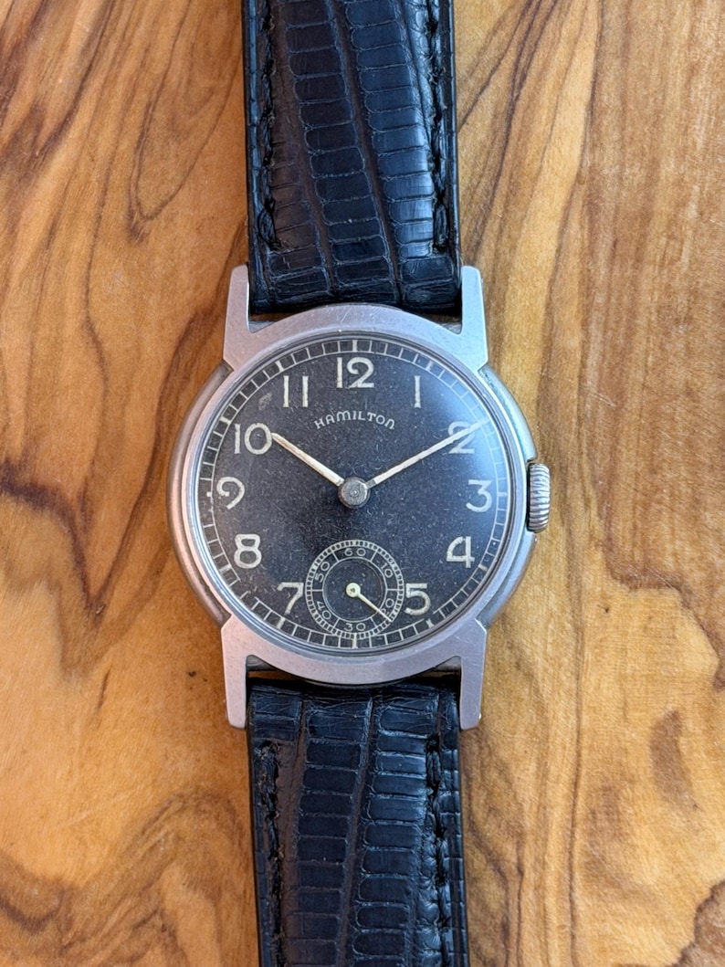 May include: A vintage Hamilton wristwatch with a black face, silver hands, and Arabic numerals. The watch has a small seconds dial and is set in a silver case with a black leather strap.