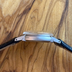 May include: A silver wristwatch with a clear, domed crystal and a black leather strap. The watch case has a rectangular shape. The watch is seen from the side, showcasing its profile against a wooden background.