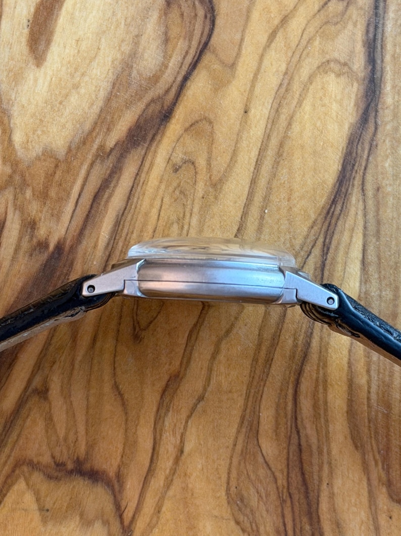 May include: A silver wristwatch with a clear, domed crystal and a black leather strap. The watch case has a rectangular shape. The watch is seen from the side, showcasing its profile against a wooden background.