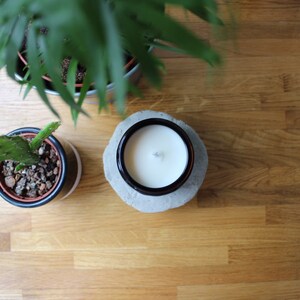 May include: A white candle with a wick in a black glass jar on a gray concrete base. The candle is on a wooden surface with two potted plants.