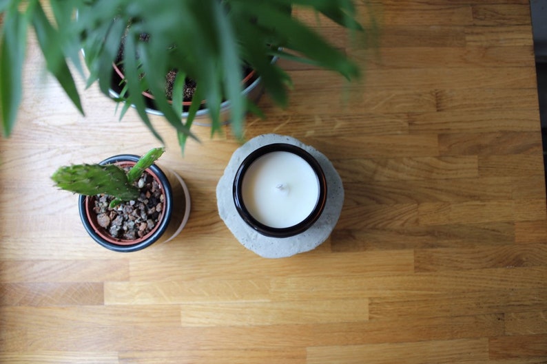 May include: A white candle with a wick in a black glass jar on a gray concrete base. The candle is on a wooden surface with two potted plants.