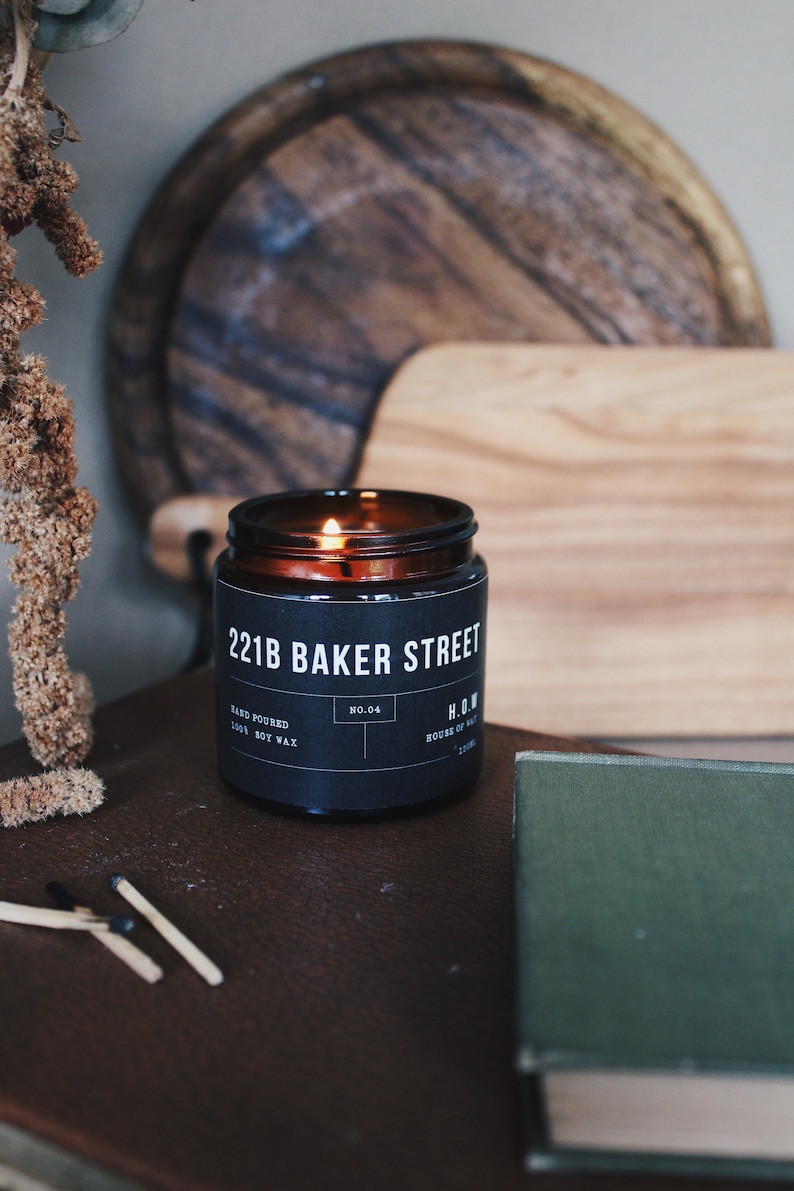 May include: A brown glass candle jar with a black label that reads "221B Baker Street" and "H.O.W. House of Wax". The candle is lit and the flame is visible.
