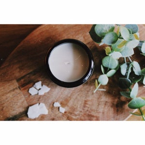 May include: A white candle in a black glass jar on a wooden surface with green leaves and white pieces of wax.