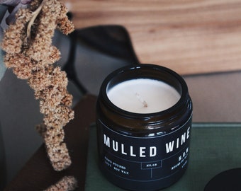 May include: A black glass candle jar with a white label that reads "Mulled Wine" and "Hand Poured Soy Wax". The candle is on a green book with a black lid.