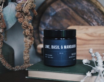 May include: A black candle jar with a white label that reads "Lime, Basil & Mandarin", "Hand Poured Soy Wax", "No. 10", and "H.O.W. House of Wax". The candle is sitting on a stack of books.