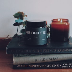 May include: Two candles, one black and one amber, are sitting on a stack of three books. The black candle has a label that reads "221B Baker Street". The books are "The Catcher in the Rye", "Julius Caesar", and "An Unkindness of Ravens".