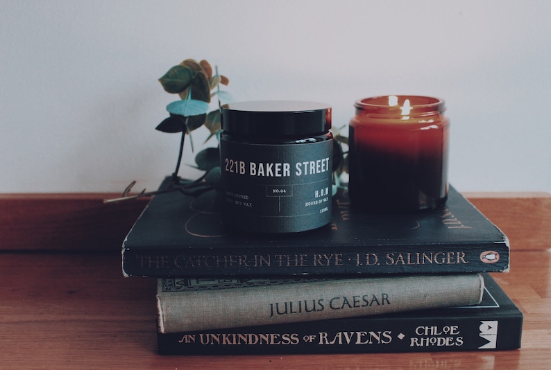 May include: Two candles, one black and one amber, are sitting on a stack of three books. The black candle has a label that reads "221B Baker Street". The books are "The Catcher in the Rye", "Julius Caesar", and "An Unkindness of Ravens".