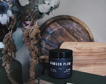 May include: A black candle jar with the label "Damson Plum" and the text "H.O.W. House of Wax 120ml" on a green book with burnt matches on a dark wooden surface.