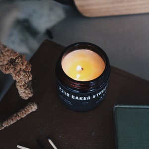 May include: A black candle jar with a yellow wax candle burning inside. The jar has the text "221B BAKER STREET" printed on the label.