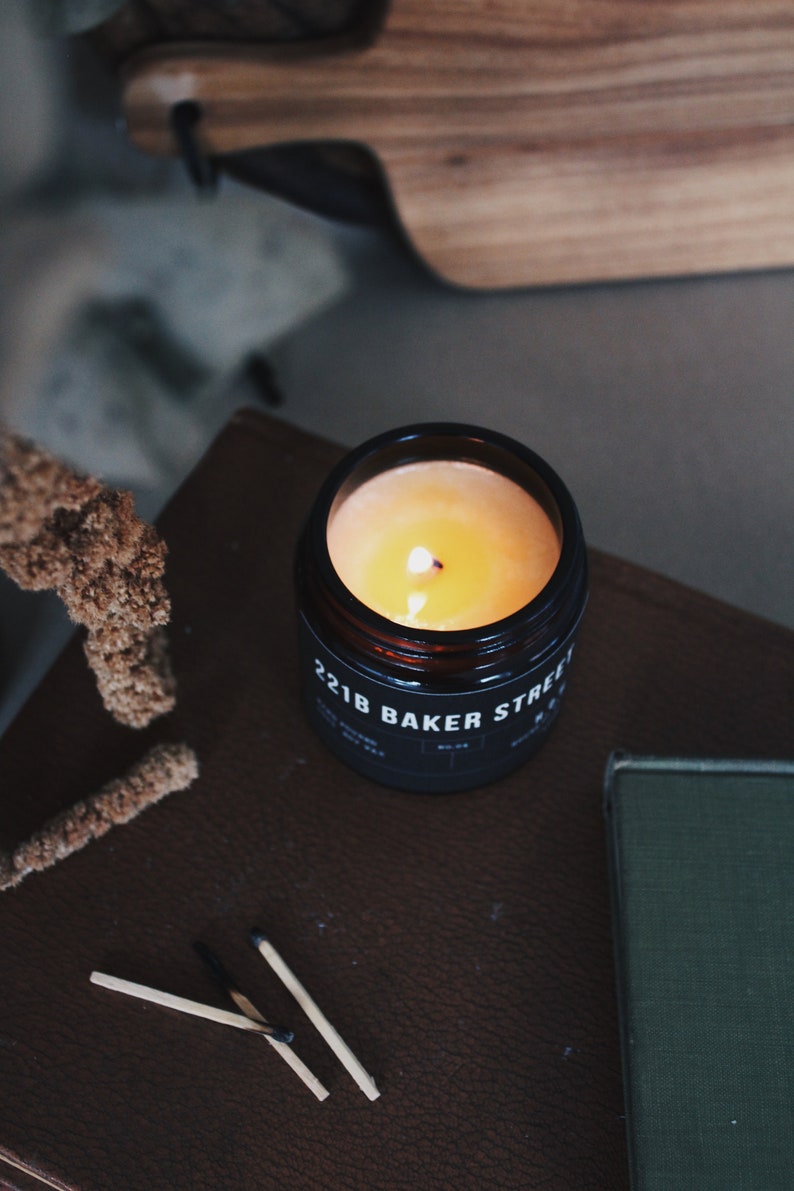 May include: A black candle jar with a yellow wax candle burning inside. The jar has the text "221B BAKER STREET" printed on the label.