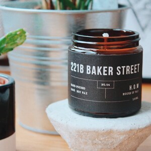 May include: A brown glass candle jar with a black label that reads "221B Baker Street" and "H.O.W. House of Wax". The candle is on a gray concrete pedestal.