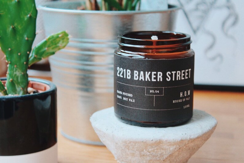 May include: A brown glass candle jar with a black label that reads "221B Baker Street" and "H.O.W. House of Wax". The candle is on a gray concrete pedestal.