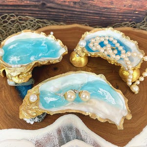 May include: Three decorative oyster shell jewelry dishes. Each shell is painted with a blue and white ocean design and accented with gold leaf. One holds a pearl necklace, another has pearl earrings, and the third has a single pearl.