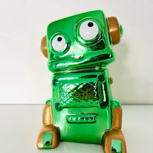 Op de afbeelding: Een metallic groene robotfiguur met gouden accenten. De robot heeft grote, ronde ogen met witte pupillen en gouden oorachtige structuren. Het lichaam is glanzend, reflecterend groen, met gouden voeten en details. De robot zit.
