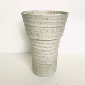 May include: A white ceramic vase with a textured, speckled finish. The vase has a wide, flared opening and a cylindrical base.