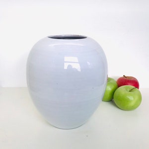 Puede incluir: Un jarrón grande de cerámica azul claro con forma redondeada. El jarrón tiene una abertura ancha y un acabado liso y brillante. Tres manzanas, dos verdes y una roja, están junto al jarrón.