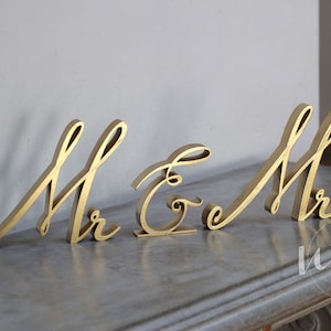 May include: Gold-painted wooden letters spelling "Mr & Mrs" in a cursive font. The letters are arranged in a line on a gray surface.