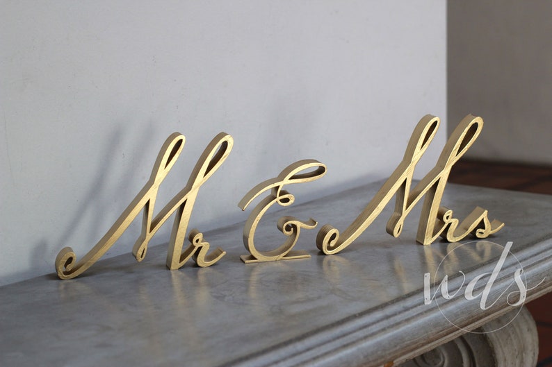 May include: Gold-painted wooden letters spelling "Mr & Mrs" in a cursive font. The letters are arranged in a line on a gray surface.
