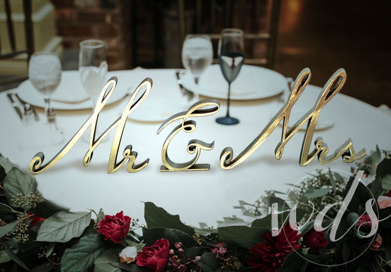 May include: Gold cursive script letters spelling "Mr & Mrs" on a white table with greenery and red flowers.