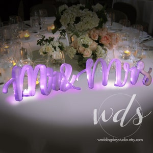 May include: Purple illuminated "Mr & Mrs" sign on a white tablecloth with a floral centerpiece and candles. The sign is made of acrylic and has a modern, elegant design.