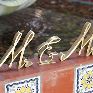 May include: Gold-painted wooden letters spelling "Mr & Mrs" in a cursive font. The letters are arranged on a tiled surface.