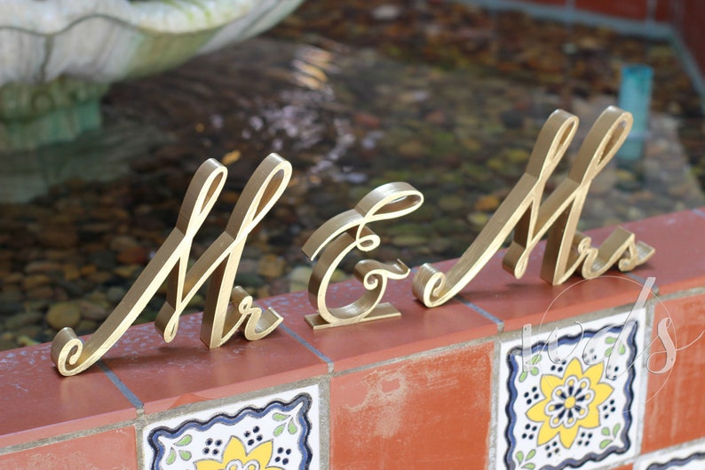 May include: Gold-painted wooden letters spelling "Mr & Mrs" in a cursive font. The letters are arranged on a tiled surface.