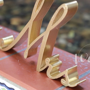May include: Gold-colored metal letters spelling "Mr." in a cursive font. The letters are standing on a brick surface.