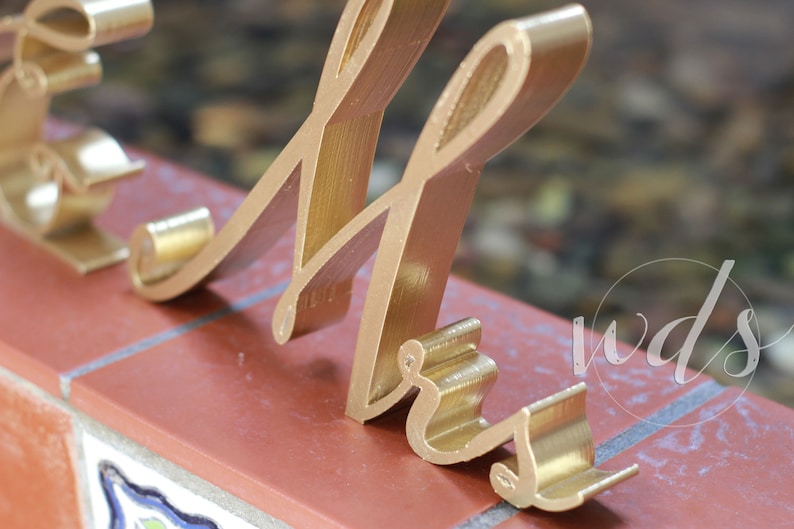 May include: Gold-colored metal letters spelling "Mr." in a cursive font. The letters are standing on a brick surface.
