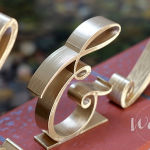 May include: Gold-painted metal letters spelling out the word "and" in a decorative script font. The letters are standing on a red brick surface.