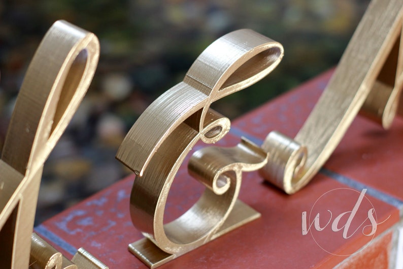 May include: Gold-painted metal letters spelling out the word "and" in a decorative script font. The letters are standing on a red brick surface.