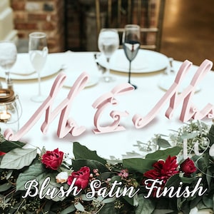 May include: Pink wooden "Mr & Mrs" signs with a blush satin finish, placed on a white table with greenery and red flowers.