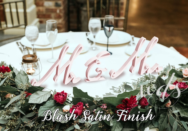 May include: Pink wooden "Mr & Mrs" signs with a blush satin finish, placed on a white table with greenery and red flowers.