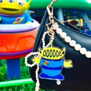 May include: A black handbag with a pearl strap and a charm featuring a cartoon alien character. The charm is yellow, blue, and clear, with the word "000000" printed on its head. The background shows a theme park ride.