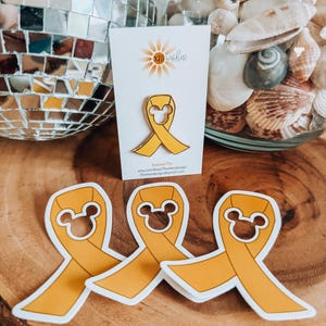May include: Three yellow enamel pins shaped like a ribbon with Mickey Mouse ears. The pins are on a wooden surface. The text on the card reads "Enamel Pin" and "etsy.com/shop/TheBesideDesign".