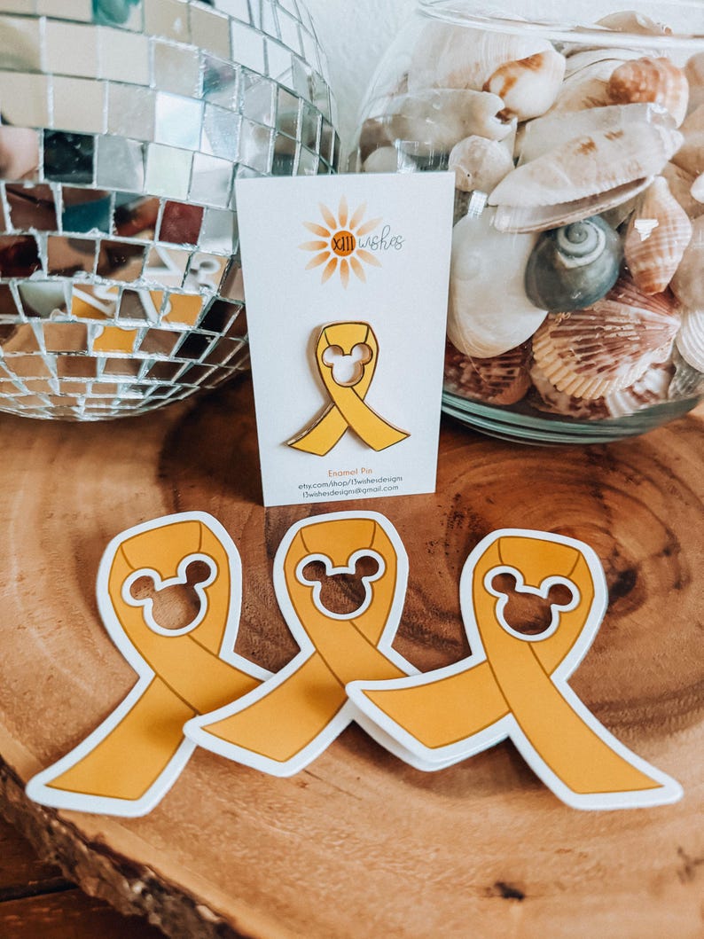 May include: Three yellow enamel pins shaped like a ribbon with Mickey Mouse ears. The pins are on a wooden surface. The text on the card reads "Enamel Pin" and "etsy.com/shop/TheBesideDesign".