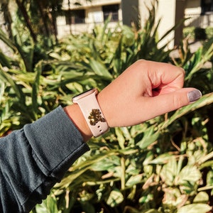 May include: A pink Apple Watch with a gold charm attached to the band. The watch is on a person's wrist.