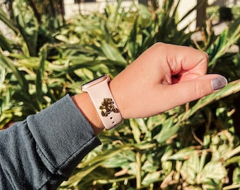 Puede incluir: Un Apple Watch rosa con un dije dorado sujeto a la banda. El reloj está en la muñeca de una persona.